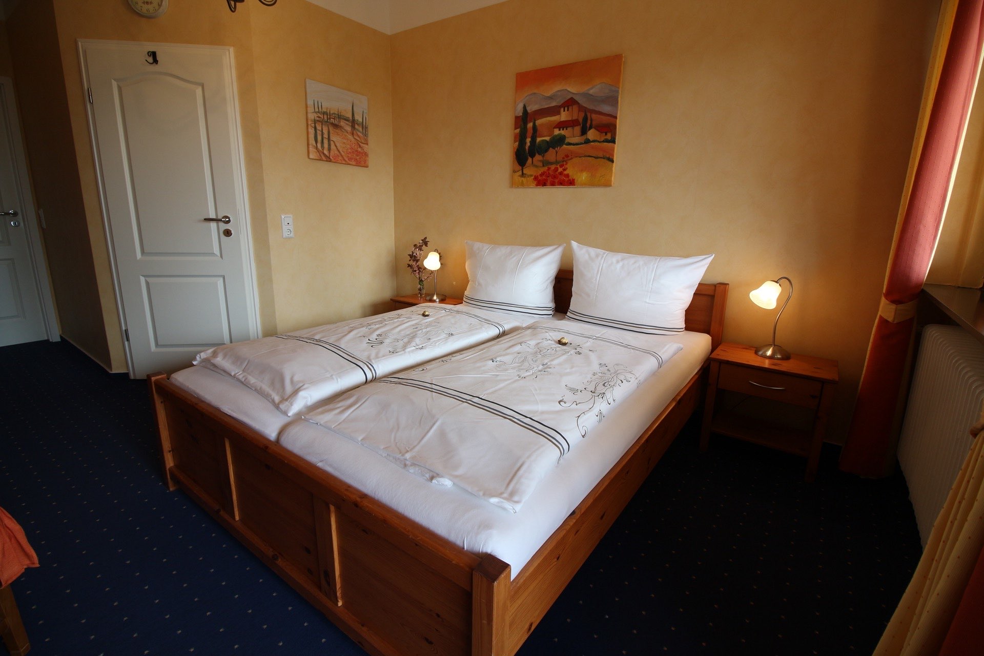 Zimmer ‚Firence‘ in der Pension Rehwinkel in Nalbach, mit gemütlichem Holzbett, warmen Gelbtönen an den Wänden, künstlerischen Landschaftsgemälden und einladender Beleuchtung.